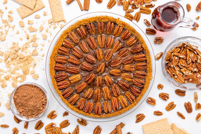 5D4B3736 - Healthy Chocolate Pecan Pie - baked pie on a white table surrounded by cinnamon, walnuts, graham crackers and maple syrup