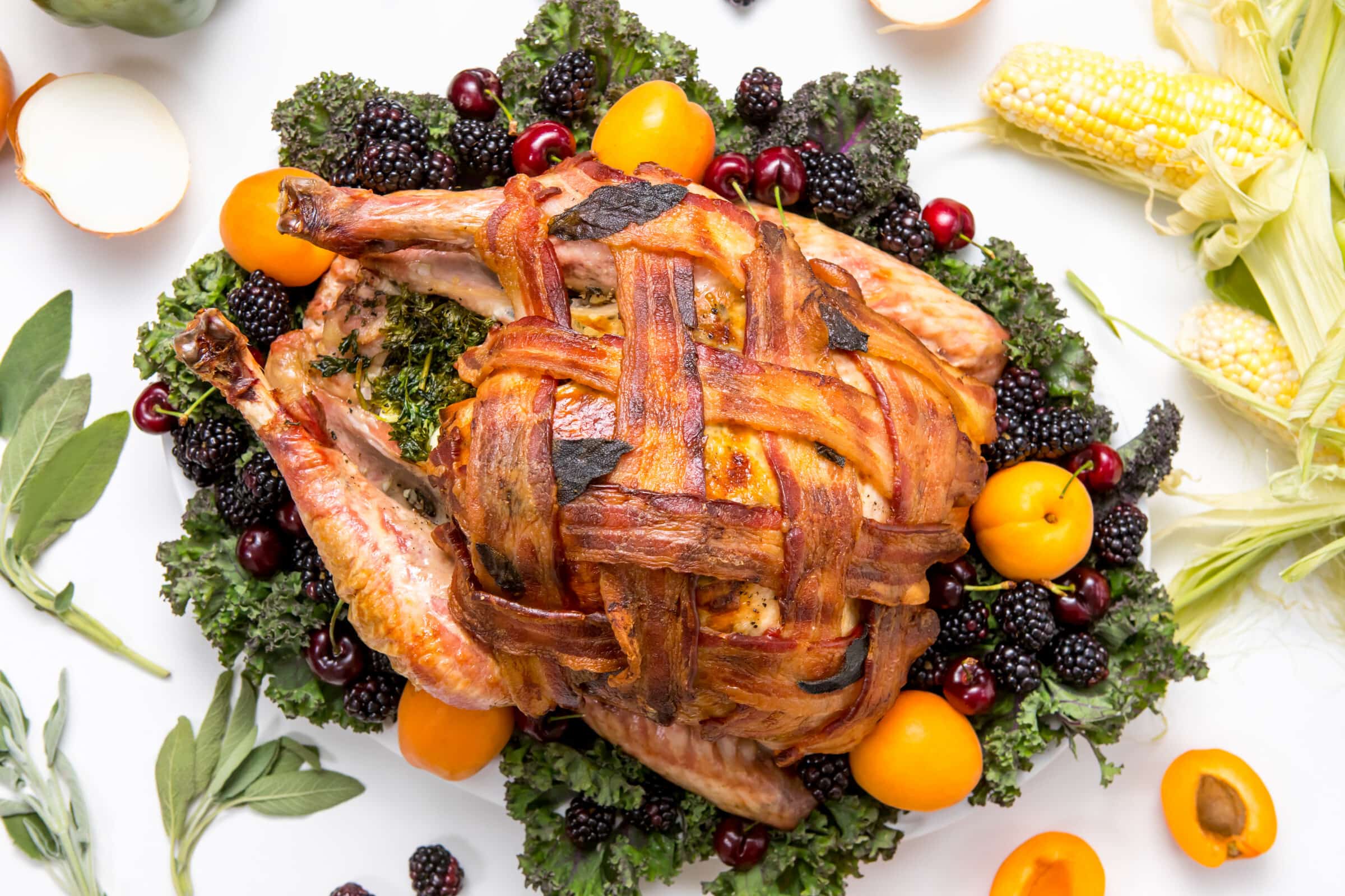 5D4B9754 - Bacon Wrapped Turkey - plated bacon wrapped turkey on a white table surrounded by corn, oranges, onions, bell pepper, blueberries, apricots and sage