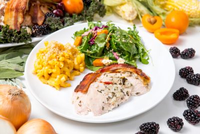 5D4B9820 - Bacon Wrapped Turkey - plated bacon wrapped turkey on a white table surrounded by corn, oranges, onions, bell pepper, blueberries, apricots and sage