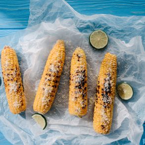 elevated view of grilled salted corn with lime slices on baking paper on wooden table